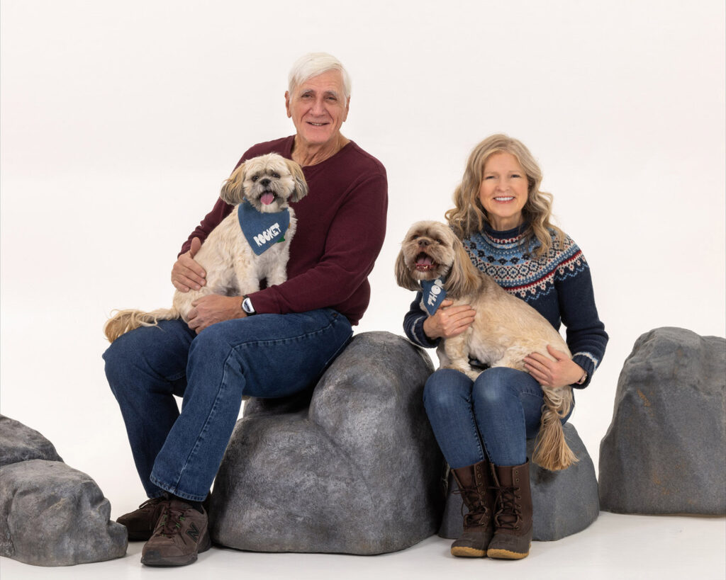A man and woman sit on rocks, enjoying the outdoors with their two dogs beside them, surrounded by nature.