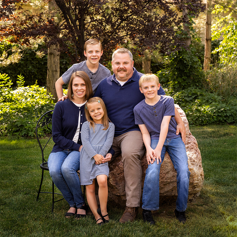 A family stands together in a park, smiling for a photo, surrounded by greenery and trees in the background.